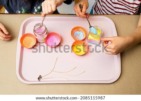 Young children making fun colorful layered granulated wax candles in home by pouring powder in old baby food jar and inserting wick inside. Hobby concept. Royalty-Free Stock Photo #2085115987