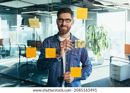 focused and thinking businessman writing on colorful notes attached to a glass wall, the entrepreneur creates a visual to-do list, business goals for future careers, daily goals, the concept of  tasks