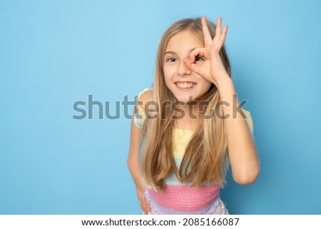 Funny little girl in summer clothing showing okay sign on eye isolated over blue background.