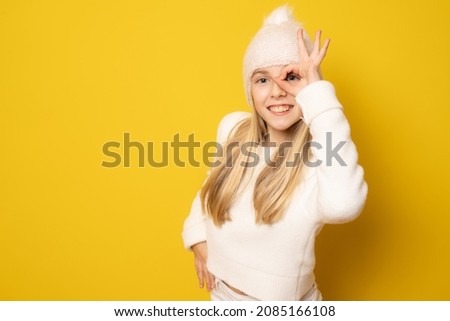 Funny little girl in winter clothing showing okay sign on eye isolated over yellow background.