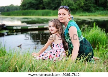 Beauty kid girl and her young mother sitting near the river on the beauty summer evening