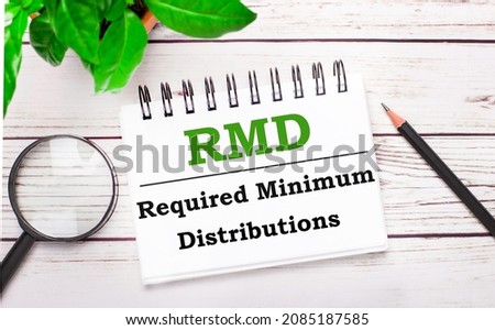 On a light wooden background, a magnifying glass, a pencil, a green plant and a white notebook with text RMD Required Minimum Distributions. Business concept