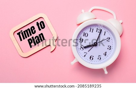 On a delicate pink background, a white alarm clock and a wooden frame with the text TIME TO PLAN