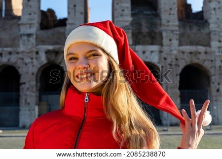 Roman holidays at Christmas. Beautiful blonde woman posing in front of the Colosseum with Santa Claus hat. The young woman looks into the camera and smiles.