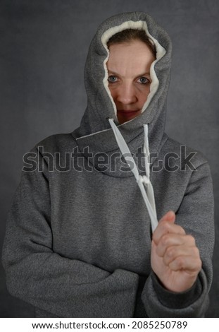 An attractive adult woman in a warm hoodie tightens the drawstrings of the hood. Photo on the background of a gray concrete wall.