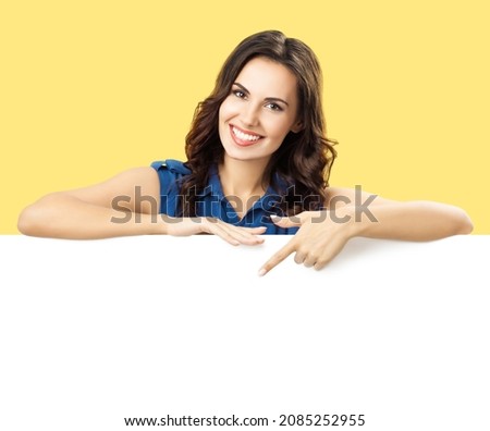 Happy smiling brunette woman in blue dress, showing, pointing, standing behind, peeping from blank banner mock up white billboard sign board signboard with ad copy space, over yellow color background. Royalty-Free Stock Photo #2085252955