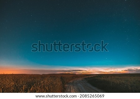 Night Starry Sky With Glowing Stars Above Countryside Landscape. Light Cloudiness Overcast Above Rural Field Meadow And Country Road In Summer.