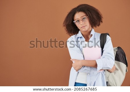 Portrait of intelligent teenage girl in glasses standing with backpack against brown background