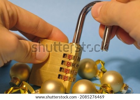 Concept. New Year. Lockdown. Blue background. Close-up. Hands hold an open combination lock with the numbers 2022. Nearby are golden Christmas tree decorations.