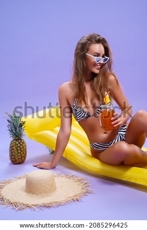  cheerful amazing beautiful caucasian woman who wears in bikini sits on inflatable yellow matress with fruit cocktail on purple background . Summer concept. Travel concept. Relax concept