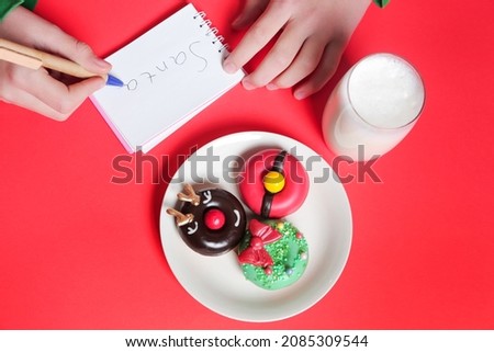 Child writes a letter for Santa, Donuts cookies and glass of milk on red background, concept Christmas and holiday.