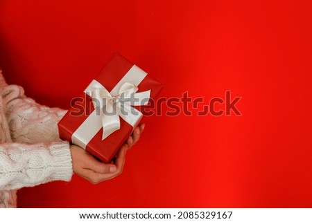 Female wearing white knitted sweater holding present tied with a silk bow over isolated red background. Christmas gift in hands of a woman. Close up, copy space for text.
