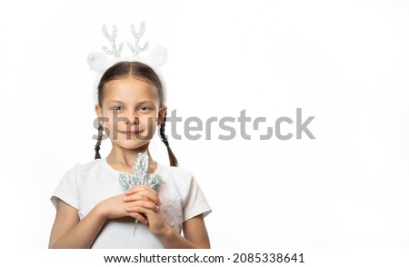 Portrait of smiling little girl in Christmas deer headband holding twig of fir tree against white background