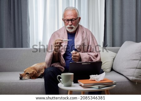 Sick elderly man checking his temperature suffering from seasonal flu or cold and using digital tablet for entertainment or online doctor consultation. Ill senior feel unhealthy with influenza at home Royalty-Free Stock Photo #2085346381