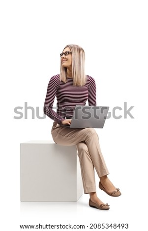 Young woman sitting on a white cube with a laptop computer and looking behind isolated on white background