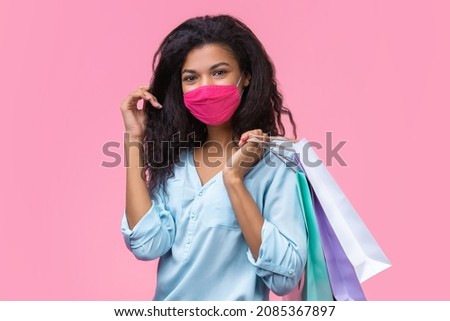 Studio shot of attractive young woman in a medical protective mask on her face, holding shopping bags isolated over pastel pink background. Safe shopping during quarantine concept.
