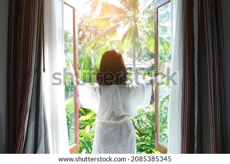 A woman opens a window to welcome the morning sunlight of a new day at her vacation home. Natural tree background.