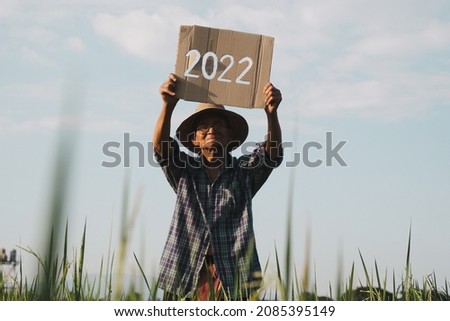 A senior farmer standing in a rice field at sunset and holding a New Year 2022 sign.