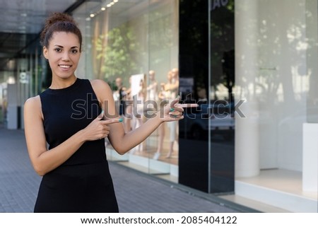advertisement, presentation, lifestyle and people concept - happy African-American woman pointing with two fingers at something and looking at the camera outdoors near the storefronts with copy space
