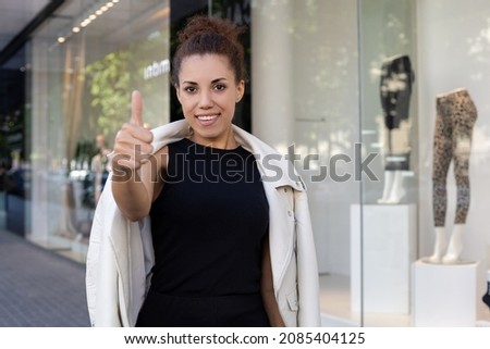 emotions, gesture, lifestyle and people concept - beautiful young african-american girl with a bundle on her head showing her thumbs up outdoors near the storefronts