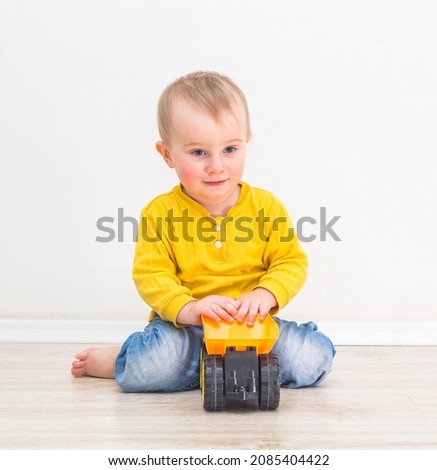One year three months old boy in yellow clothes sitting and playing with a toy truck on white wall background
