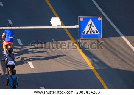 High angle view and focus at overhead electric pedestrian crossing sign over with blurred background of cyclist group riding on the road at morning time