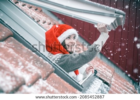 Young woman in a red and white Christmas hat opening a skylight window and smiling, excited about first snow. 