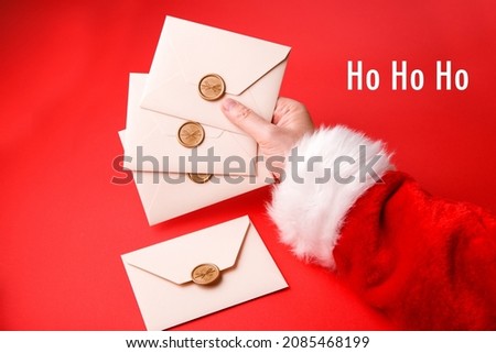 Santa Claus holds in his hand a three beige envelopes and one lies on a red background. Christmas mail for Santa Claus with an inscription Ho Ho Ho. Top view