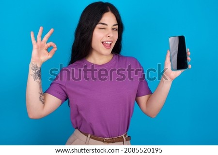 Excited Caucasian woman wearing purple T-shirt over blue background showing smartphone blank screen, blinking eye and doing ok sign with hand.  Advertisement concept.