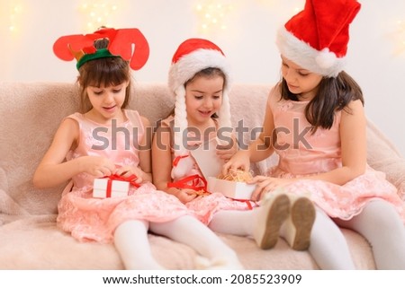 portrait of girls celebrating christmas or new year, children dressed in santa helper hat, sitting on a couch in home interior decorated with christmas lights and new year holiday gifts