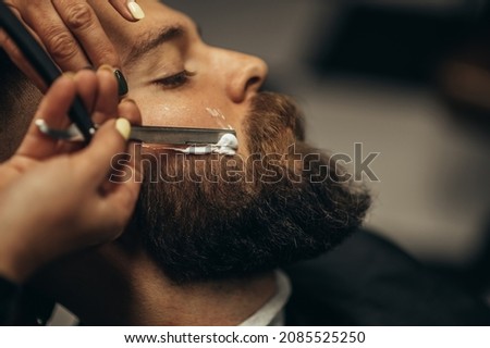 Young bearded man getting shaved with straight edge razor by hairdresser at barbershop Royalty-Free Stock Photo #2085525250