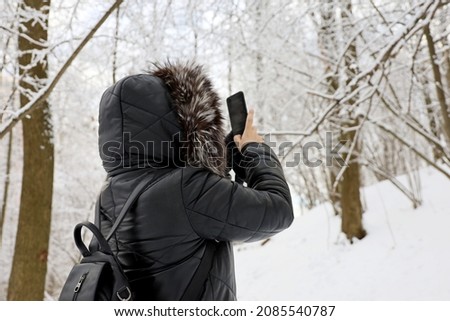 Woman in black coat with fur hood taking pictures of snow nature on a smartphone in the winter forest. Trees after snowfall, leisure at cold weather