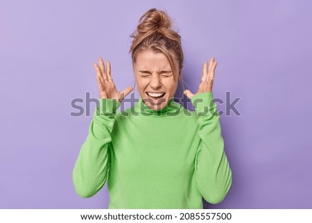 Emotional young European woman screams loudly keeps hands raised near head expresses anger being fed up of something wears casual green jumper isolated over purple background. Human emotions concept Royalty-Free Stock Photo #2085557500