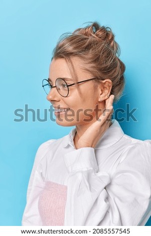 Profile shot of happy female student looks away joyfully rests after lectures wears round spectacles formal white shirt isolated over blue studio wall. Intelligent woman comes on job interview