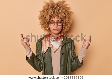 Calm relaxed young curly woman gathers patience makes mudra sign breathes deeply keeps eyes closed wears leather shirt kerchief tied around neck reaches nirvana isolated over beige background