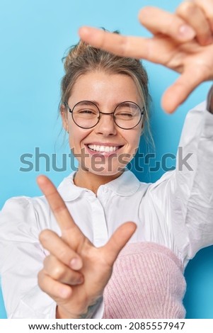 Young positive lovely woman looks through hands frame winks eye and smiles pleasantly imagines something interesting searches for photo angle poses against blue background takes picture of moment