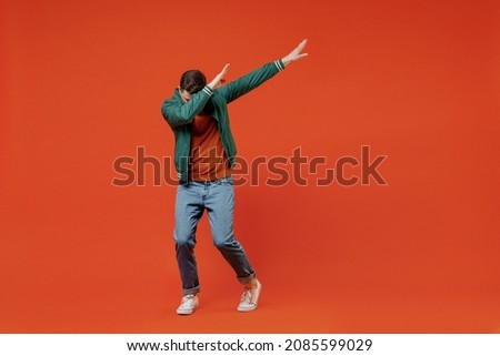 Full size body length fun young brunet man 20s wears red t-shirt green jacket doing dab hip hop dance hands move gesture youth sign hide cover face isolated on plain orange background studio portrait.
