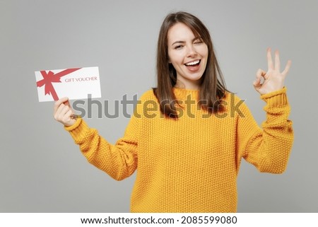 Young smiling positive attractive caucasian woman in casual knitted yellow sweater point index finger on gift voucher flyer mock up show ok okay gesture isolated on grey background studio portrait.