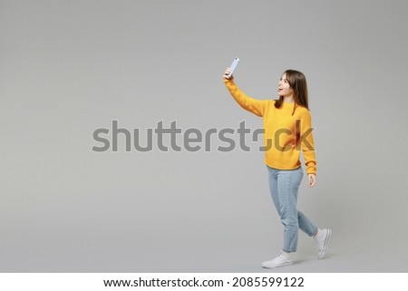 Full length body of young smiling student positive caucasian happy woman 20s wearing casual knitted yellow sweater doing selfie shot on mobile phone isolated on grey color background studio portrait.