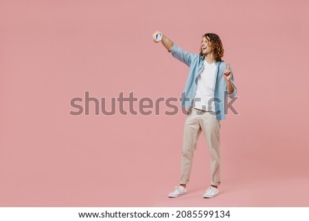 Full length young happy man with long curly hair wear blue shirt white t-shirt doing selfie shot on mobile cell phone post photo on social network isolated on pastel plain pink wall background studio.