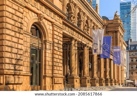 historic building of the stock exchange in Frankfurt with flags above the entrance. brown colored commercial building in baroque style. Skyscrapers in the background. Trees in the foreground in spring