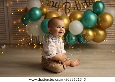 Cute blond kid in a festive shirt and trousers. A happy kid on the background of balloons with garlands celebrates his birthday. High-quality photography