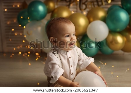 Cute blond kid in a festive shirt and trousers. A happy kid on the background of balloons with garlands celebrates his birthday. High-quality photography