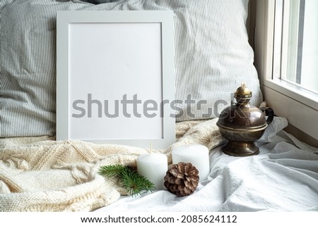 Cozy winter home interior scene.Christmas composition near the window.Blank white picture frame mockup, oil lamp, candles,pine cone.Model for Christmas menu.