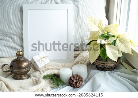Cozy winter home interior scene.Christmas composition near the window.Blank white pictur frame mockup, white poinsettia, oil lamp, candles, gift box.Model for Christmas menu.