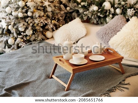 Picture of wooden tray with teapot, cups on grey bed in living room with Christmas decoration over new year tree background. Nordic scandinavian country cottage style. Christmas card concept
