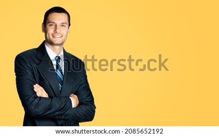 Portrait of happy smiling brunette businessman in black suit, white shirt and blue stripes tie standing in crossed arms pose over orange yellow color background. Confident business man. Male executive