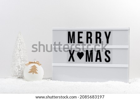 White Christmas tree, Christmas bauble and lightbox with text MERRY XMAS on a table with snow. Minimal Christmas or New Year concept.