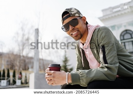 People, technology, travel and tourism - man with earphones, smartphone and bag sitting on city street bench and listening to music while drinking coffee