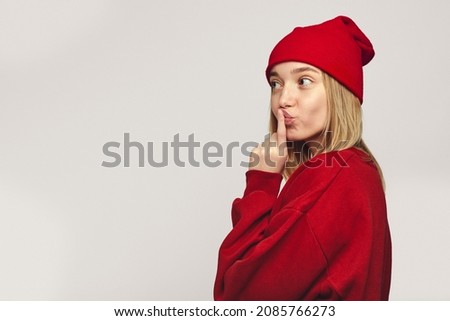 Horizontal shot of hipster girl, kissing index finger, looks at empty free space, wears stylish red outfit, isolated over white background
